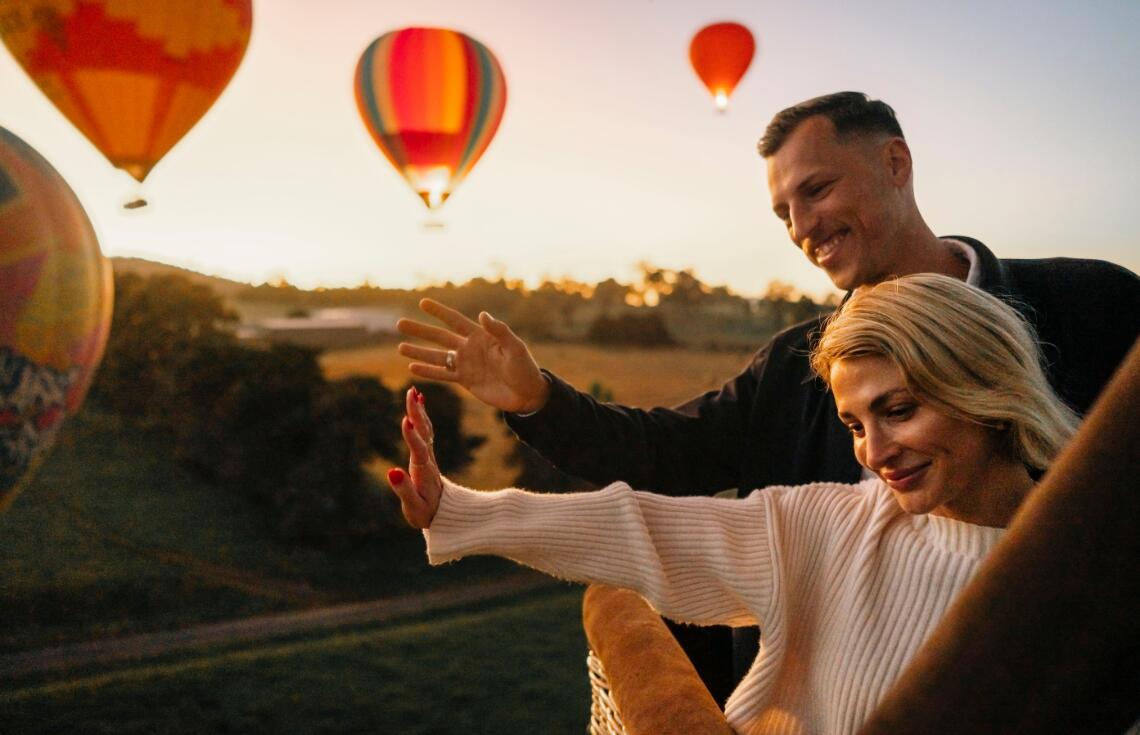 A couple waving to the ground as they lift off in a balloon basket with hot air balloons in the background.