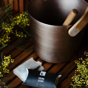 A sauna bucket surrounded by native flora and a sauna scent packet with birch in the foreground.